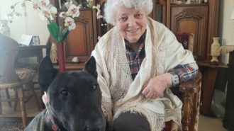 Foto: Ältere Seniorin mit weißen Haaren sitzt mit ihrem Hund in ihrem Wohnzimmer und sieht freundlich in die Kamera.